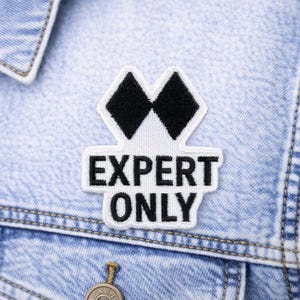May include: A white and black patch with the words "EXPERT ONLY" is sewn onto a light blue denim jacket. The patch features two black diamond shapes above the text.