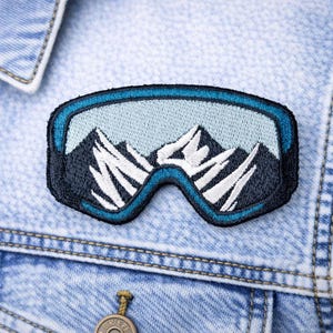 May include: Embroidered patch depicting ski goggles with a mountain range scene. The patch features a blue border and a light blue lens area, with white and black mountain peaks. The patch is sewn onto a light blue denim jacket.