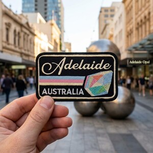 May include: A black rectangular patch with the word "Adelaide" in white script above "AUSTRALIA" in white capital letters. The patch features a colorful geometric design and a horizontal stripe design in pink, blue, and yellow.