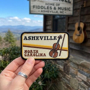 May include: A rectangular embroidered patch with a cream background and black border. It features the text 'ASHEVILLE' and 'NORTH CAROLINA' in brown, alongside a brown violin and bow graphic, with a horizontal gold stripe across the center.
