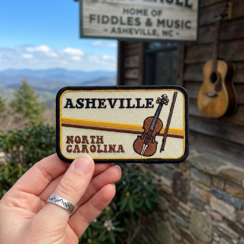 May include: A rectangular embroidered patch with a cream background and black border. It features the text 'ASHEVILLE' and 'NORTH CAROLINA' in brown, alongside a brown violin and bow graphic, with a horizontal gold stripe across the center.