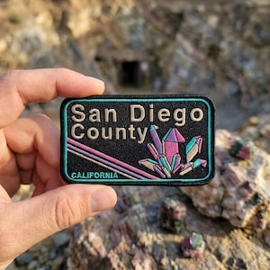 May include: A rectangular embroidered patch with a black background and a teal border. The text reads San Diego County in beige and CALIFORNIA in teal. A cluster of multi-colored crystals and three diagonal lines in pink and purple are featured.