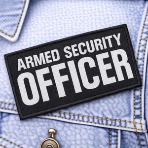 May include: A black rectangular patch with the words "ARMED SECURITY OFFICER" in bold white letters. The patch is sewn onto a light blue denim jacket. The jacket has visible stitching and a metal button.