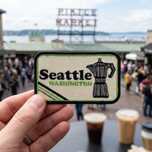 May include: A rectangular embroidered patch with a cream-colored background and a black border. It features the text 'Seattle' in black and 'WASHINGTON' in green, alongside a black and white illustration of a moka pot coffee maker.