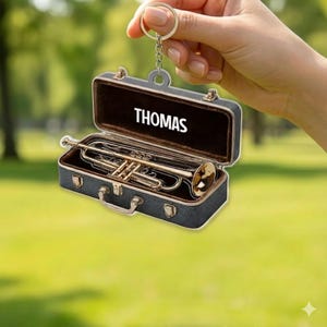 May include: A miniature trumpet keychain inside a black case with the name "THOMAS" in white letters. The trumpet is gold-colored, and the case is open, revealing a brown interior. The keychain is held by a hand against a blurred green background.