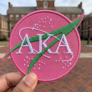 May include: A pink embroidered patch with the letters "AKA" in white, overlaid with a green swoosh design. The patch is circular and features a NASA-inspired design with white stars. The patch is held in front of a brick building.