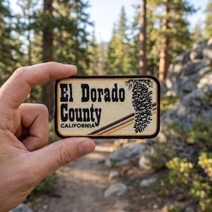 May include: A rectangular embroidered patch with a cream-colored background and a black border. The text reads El Dorado County CALIFORNIA in black lettering. A black pinecone graphic and two diagonal brown stripes are on the right side.