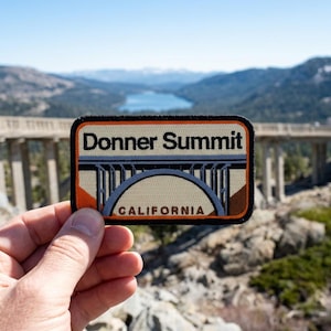 May include: A rectangular embroidered patch featuring a graphic of the Donner Summit bridge in California. The design includes the text Donner Summit and California in black lettering against a cream, orange, and grey background with a black border.