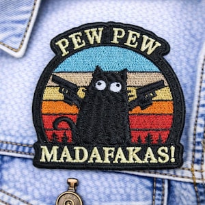 May include: Embroidered patch featuring a black cat holding two guns, with the text "PEW PEW MADAFAKAS!". The patch has a retro sunset design with blue, yellow, orange, and red colors. It is attached to a denim jacket.