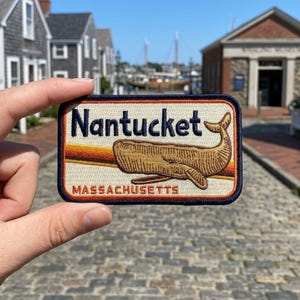 May include: A rectangular embroidered patch with a dark blue border, featuring the text "Nantucket" in dark blue and "MASSACHUSETTS" in orange. A tan whale is centered over horizontal stripes of yellow, orange, and brown on a cream background.