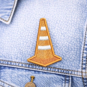 May include: Embroidered patch of an orange traffic cone with three white stripes. The patch is sewn onto a light blue denim jacket. The cone is a common symbol for road work and construction.
