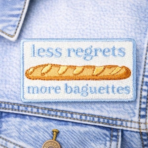 May include: Embroidered patch with the text "less regrets more baguettes" and a baguette illustration. The patch is rectangular with a light blue border and is sewn onto a light blue denim jacket.