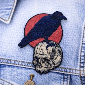 May include: Embroidered patch featuring a dark blue crow perched atop a skull, set against a red circle. The patch is sewn onto a light blue denim jacket, showcasing a gothic or alternative aesthetic.