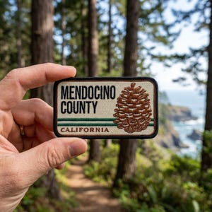 May include: A rectangular embroidered patch with a cream background and black border. It features the text MENDOCINO COUNTY in black, a brown pinecone illustration, two horizontal green stripes, and the word CALIFORNIA in brown text.
