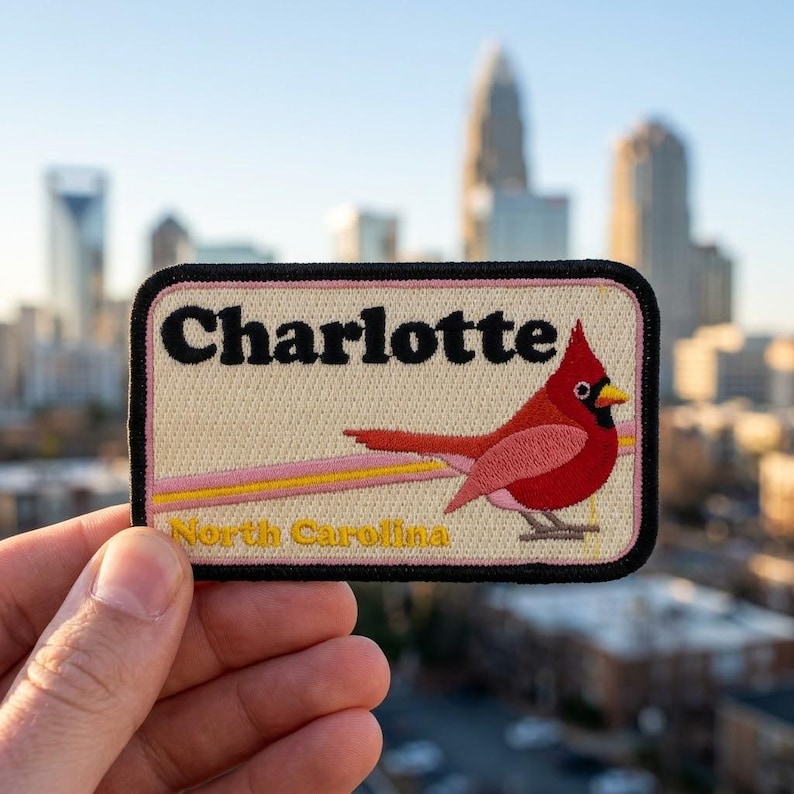 Puede incluir: Un parche rectangular bordado con fondo crema y borde negro. Presenta el texto "Charlotte" en negro y "North Carolina" en amarillo, con un p&aacute;jaro cardenal rojo posado sobre una l&iacute;nea horizontal de color rosa y amarillo.