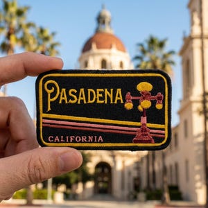 May include: A rectangular embroidered patch with a black background, featuring the yellow text PASADENA and CALIFORNIA. It includes a yellow and pink striped design and a graphic of a yellow and red street lamp, all bordered by a yellow edge.