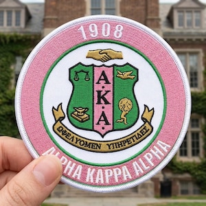 May include: A round embroidered patch with a pink border and white center. The patch features the text "1908" at the top and "ALPHA KAPPA ALPHA" at the bottom. The center design includes a green shield with gold and pink accents.