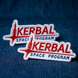 May include: Two embroidered patches with the text "KERBAL SPACE PROGRAM" in red and blue on a white background. The patches feature a stylized rocket design and are set against a blue denim background.