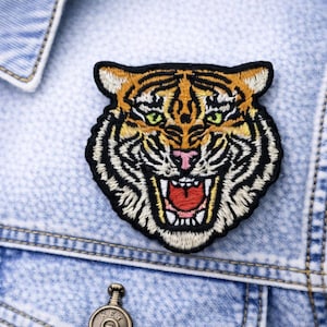 May include: Embroidered tiger head patch with orange, black, and white details, featuring a snarling expression and green eyes. The patch is affixed to a light blue denim jacket, showcasing a classic style.
