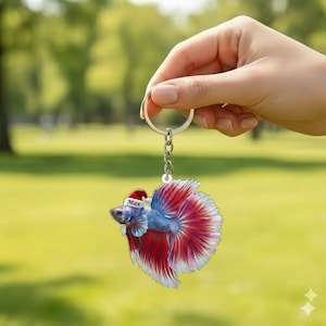 May include: A keychain featuring a betta fish design with blue and red coloring, wearing a Santa hat. The keychain is held by a hand against a blurred green background. The name "Max" is printed on the Santa hat.