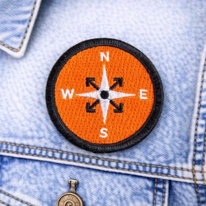 May include: Round orange compass patch with black border, stitched onto a denim jacket. The compass rose features white lettering for the cardinal directions and black directional arrows. A stylish accessory for jackets or bags.