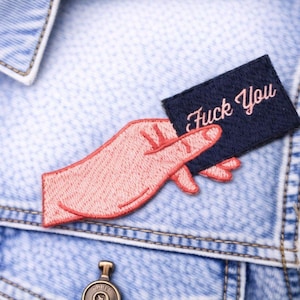 May include: Embroidered patch featuring a hand giving the middle finger, holding a navy blue rectangle with the words "Fuck You" in pink script. The patch is sewn onto a light blue denim jacket.