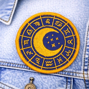 May include: A round, embroidered patch featuring a crescent moon and stars in gold against a dark blue background. The patch is encircled by zodiac symbols in gold, set against a dark blue border, and is sewn onto a denim jacket.