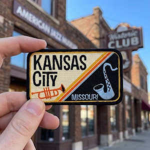 May include: A rectangular embroidered patch featuring the text Kansas City Missouri. The design is split diagonally with a cream background on the left showing an orange trumpet and a black background on the right showing a white saxophone.