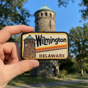 May include: Embroidered patch featuring a brown tower with a green dome, the word "Wilmington" in gold lettering, and "Delaware" in white. The patch has a black border and a retro design.