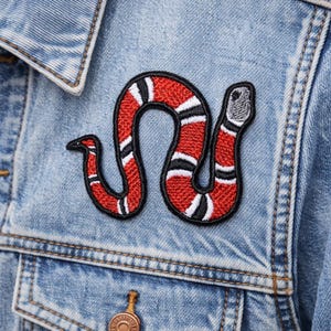 May include: A red, black, and white snake patch is sewn onto a light blue denim jacket. The snake design is detailed with a black outline and white and black stripes. The jacket has a button closure and pockets.
