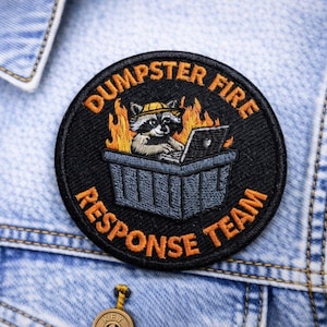 May include: Embroidered patch with the words "Dumpster Fire Response Team" in orange on a black background. The patch features a raccoon wearing a hard hat and working on a laptop inside a dumpster with flames.