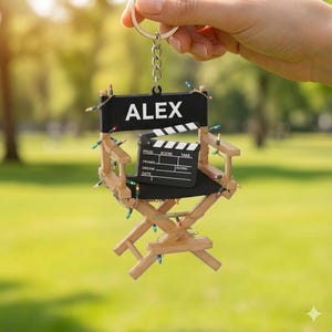 May include: A miniature director's chair keychain with the name "ALEX" on the backrest. A small film clapperboard sits on the chair. The chair is made of wood and black fabric, with colorful lights wrapped around it.