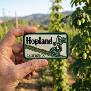 May include: A rectangular patch with the word "Hopland" in a bold, dark green font, and "CALIFORNIA" in a smaller font. The patch features a green hop plant illustration. The patch has a cream-colored background with green and brown accents.