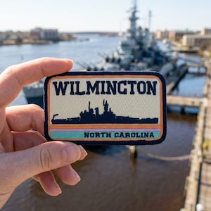 May include: A rectangular embroidered patch featuring the text WILMINGTON in dark blue, a silhouette of a battleship in dark blue, and the text NORTH CAROLINA in dark blue, with horizontal stripes in orange, pink, and light blue on a cream background.
