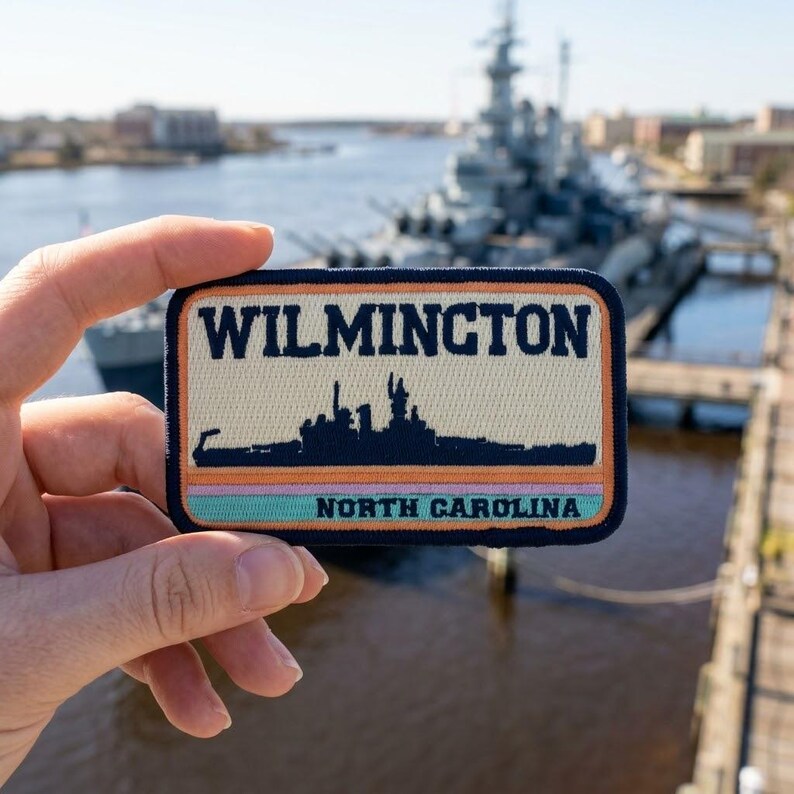 May include: A rectangular embroidered patch featuring the text WILMINGTON in dark blue, a silhouette of a battleship in dark blue, and the text NORTH CAROLINA in dark blue, with horizontal stripes in orange, pink, and light blue on a cream background.