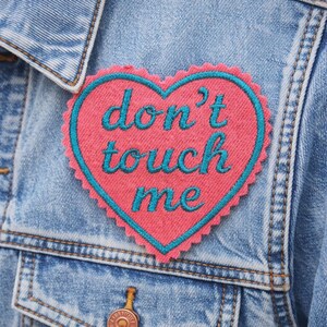May include: Close-up of a light blue denim jacket with a pink heart-shaped patch. The patch has a teal border and teal cursive text that reads "don't touch me". The jacket has a button closure and pocket.