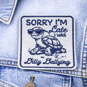 May include: A square patch with a turtle wearing sunglasses, embroidered in navy blue on a white background. The text "SORRY I'M Late I WAS Dilly Dallying" is above and below the turtle. The patch is sewn onto a denim jacket.