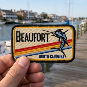 May include: Rectangular patch with the word "Beaufort" in black letters above a blue marlin illustration. The patch has a tan background with red, yellow, and blue stripes and the text "North Carolina".