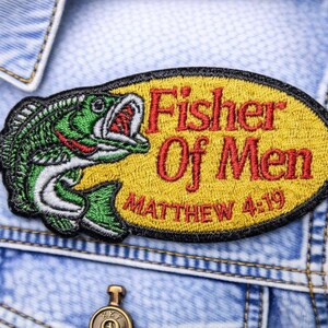 May include: Embroidered patch with a green and white fish leaping from the water, with red accents. The patch has the text "Fisher Of Men" in red, and "Matthew 4:19" in red on a yellow oval background, with a black border.