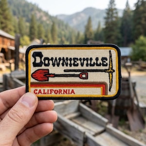 May include: Embroidered patch featuring the word "Downieville" in black letters above a red shovel and pickaxe design. The word "CALIFORNIA" is printed in red at the bottom. The patch has a tan background with a black and yellow border.