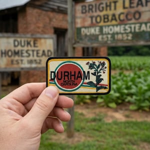 May include: A rectangular patch with the word "DURHAM" in black letters inside a red circle, with "NORTH CAROLINA" below. The patch features a floral design and is bordered in black, yellow, and green. The background includes signs for "DUKE HOMESTEAD" and "BRIGHT LEAF TOBACCO".