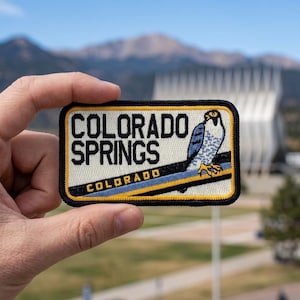 May include: A rectangular embroidered patch featuring the text COLORADO SPRINGS in black block letters, with the word COLORADO below in yellow. A blue and white falcon sits on a black and yellow diagonal stripe against a cream background.