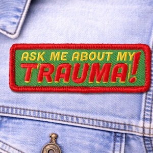 May include: Embroidered patch with the text "ASK ME ABOUT MY TRAUMA!" in red and yellow on a green background, framed by a red border. The patch is sewn onto a denim jacket.