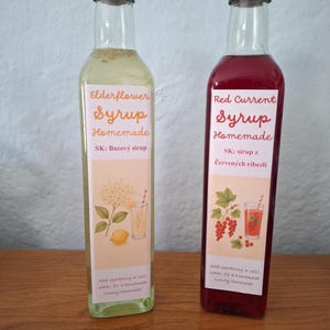 Peut inclure: Deux grandes bouteilles en verre avec des bouchons à vis noirs contenant du sirop. La bouteille de gauche contient du sirop de fleur de sureau jaune pâle, et celle de droite du sirop de groseille rouge. Chaque étiquette blanche porte du texte.