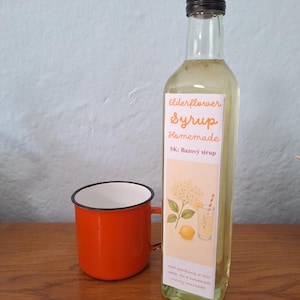 Könnte beinhalten: Eine klare Glasflasche mit hellgelbem Holunderblütensirup, schwarzem Schraubverschluss und einem Etikett mit der Aufschrift Elderflower Syrup Homemade SK: Bazový sirup, neben einem kleinen, leuchtend orangefarbenen Emaillebecher mit schwarzem Rand.