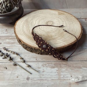 Op de afbeelding: Een bruine macramé choker ketting met goudkleurige kralen en een centrale steen. De ketting wordt getoond op een houten schijf, met gedroogde lavendel en een kleine kom op de achtergrond. De choker is ongeveer 30 cm lang.