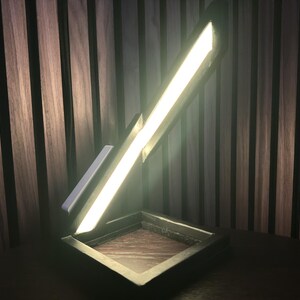 May include: Contemporary desk lamp with a rectangular base and a bright, angled light bar. The lamp has a wooden base and a metallic frame, emitting a warm glow. The background is a wooden wall.