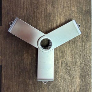 May include: A silver USB flash drive with a unique three-pronged design. The metal device has a central circular opening and is resting on a dark wooden surface. The flash drive is a modern tech accessory.