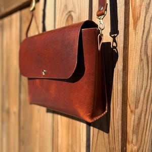 May include: A brown leather messenger bag with a flap closure and adjustable shoulder strap. The bag is hanging on a wooden fence. The leather has a rich, textured appearance, and the hardware is gold-toned.