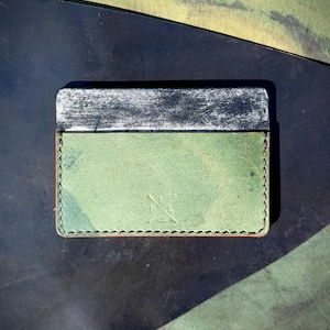 Op de afbeelding: Handgemaakte kaarthouder met een groene leren behuizing en een zilverkleurige metalen bovenkant. Het leer heeft een verweerde uitstraling met een subtiel patroon. De randen zijn gestikt met een contrasterende draad en een klein symbool is op de voorkant in reliëf aangebracht.